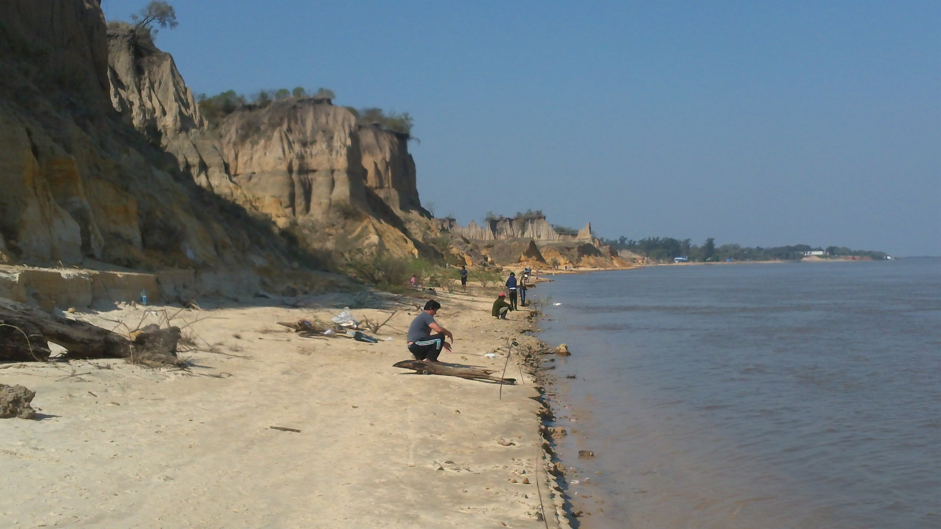 Men squatting on a river bank fishing with a sandy beach and cliff behind them.