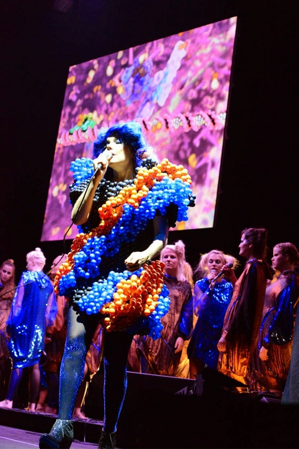 Photo of singer Björk performing dressed with a costume represening the DNA structure, with backing singers and a projection of Drew Berry showing on the background wall.