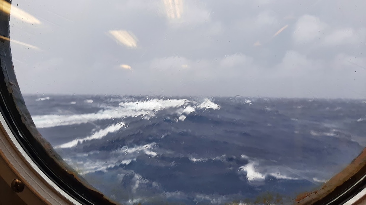 A photo out of the porthole of a boat. The focus is on the glass and out of focus through the glass the tops of crashing waves are visible  